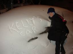 Kelly smiles impishly next to a snow drift after having just carved out the words Kelly's Car into a snow drift, which is actually her car covered in snow