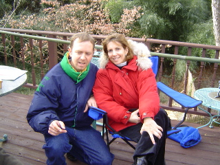 Peter kneeling beside Linda who is seated on a blue chair on the porch on a sunny winter day.